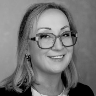 A person with glasses smiles in a black and white portrait, with shoulder-length hair and a neutral background.
