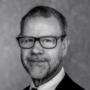 Man med glasögon och skägg ler i svartvitt porträtt.