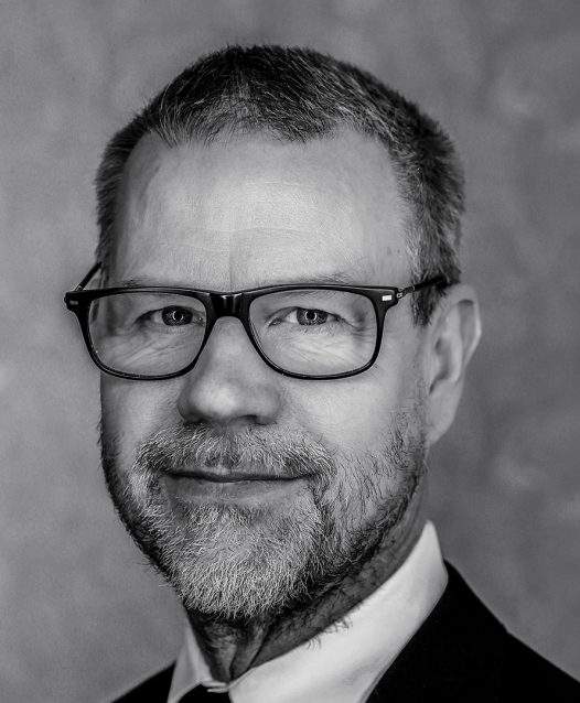 Man med glasögon och skägg ler i svartvitt porträtt.